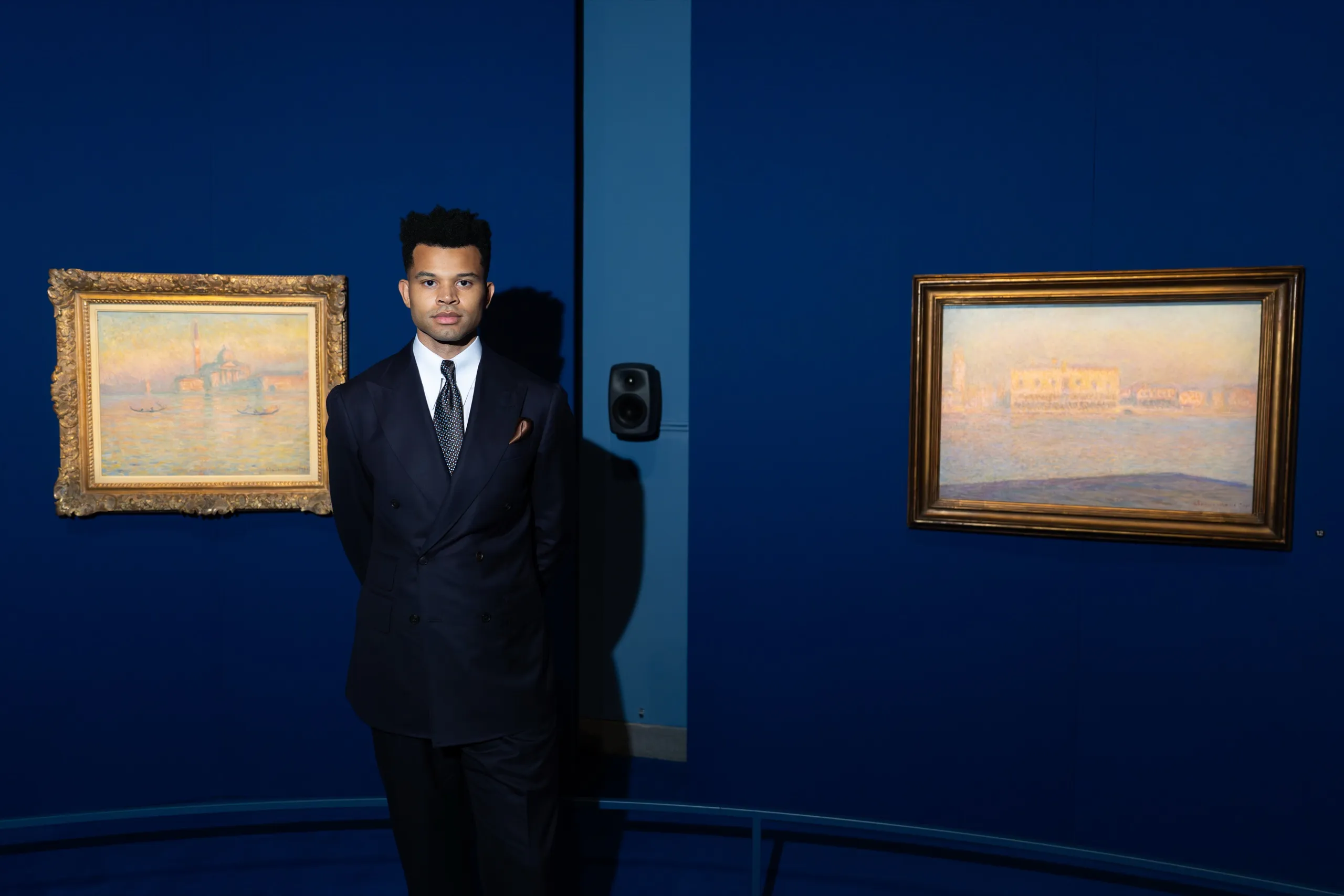 Niles Luther in the installation gallery between two Monet paintings of Venice, with a wall-mounted monitor beside him
